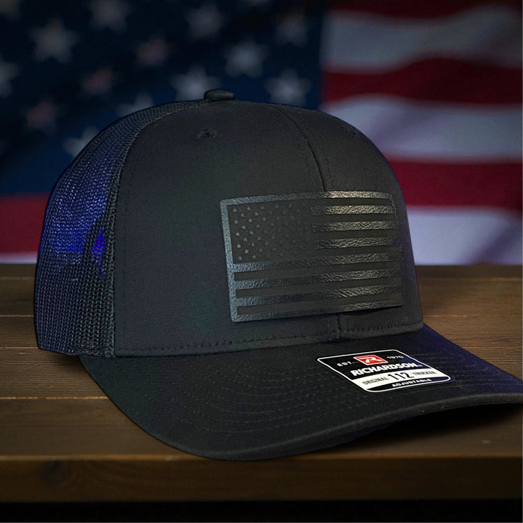 Blue trucker hat with eagle emblem and '250 Years of America' design on a wooden surface with an American flag in the background.