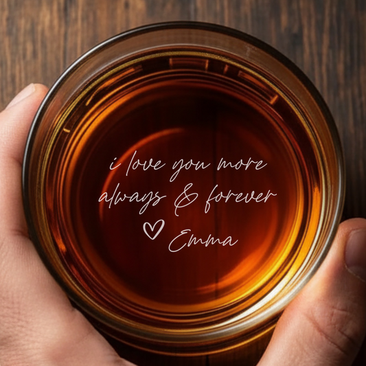 Hand holding a glass of amber liquid with 'i love you more always & forever Emma' text on a wooden surface.